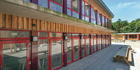 Köster verbaute bodentiefe Fenster für lichtdurchflutete Räume, die sich mit den roten Rahmen deutlich von der Fassade abheben.